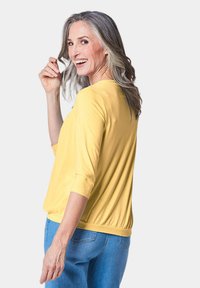Gelbes Langarmtop mit rundem Ausschnitt und gerafftem Saum; getragen mit hellblauen, engen Jeans, die eine weiche, glatte Stofftextur zeigen.