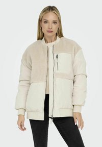 Giacca bomber beige chiaro con pannello superiore plush e testurizzato, chiusura frontale con zip, dettagli di tasche e polsini e orlo a coste.
