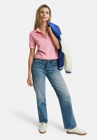 Rosa Polohemd mit kurzen Ärmeln, blaue und weiße Jacke über dem Arm, kombiniert mit hellblauen Jeans und weißen Sneakern.