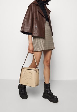 Nainen ruskea nahkatakki, musta pooloneule, ruudullinen hame, mustat korolliset saappaat ja beige Love Moschino -käsilaukku kädessään.