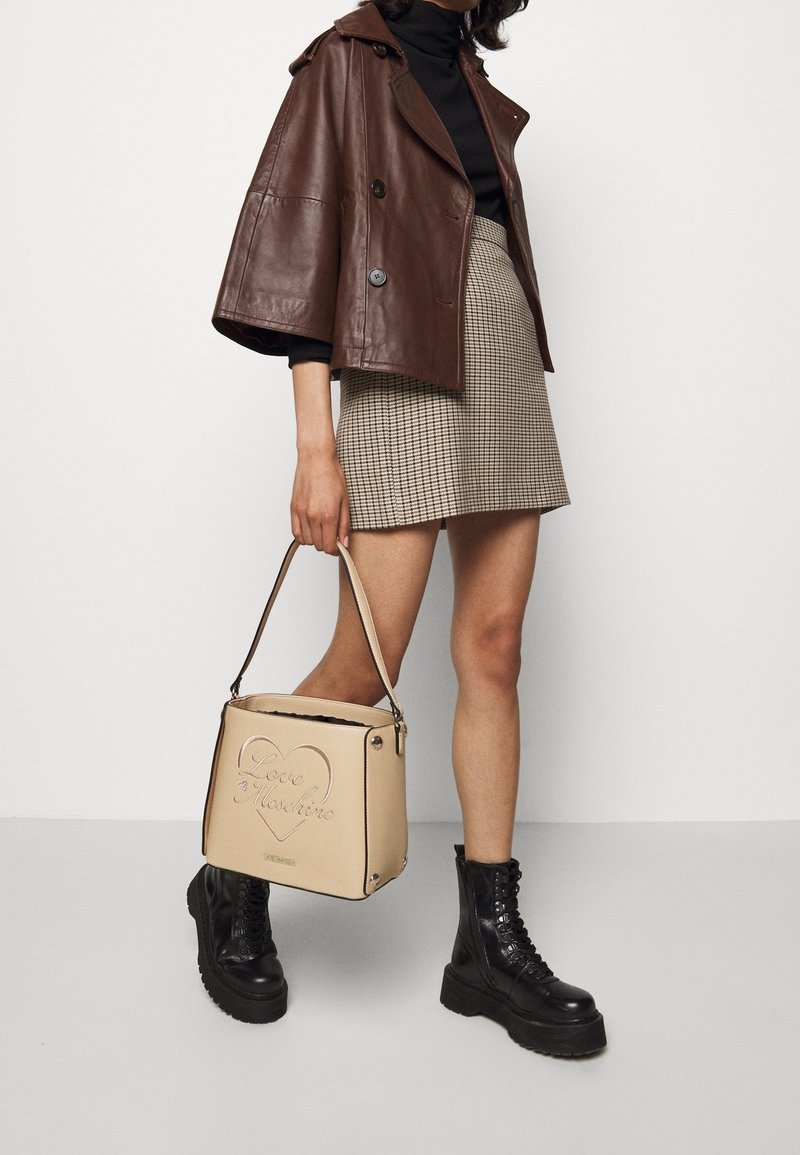 Femme portant une veste en cuir marron, un col roulé noir, une jupe à carreaux, des bottes à plateforme noires, tenant un sac à main beige Love Moschino sur le côté.