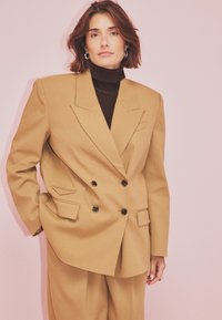 Woman with short brown hair wearing a tan oversized double-breasted blazer and pants with a black turtleneck against a pink background.