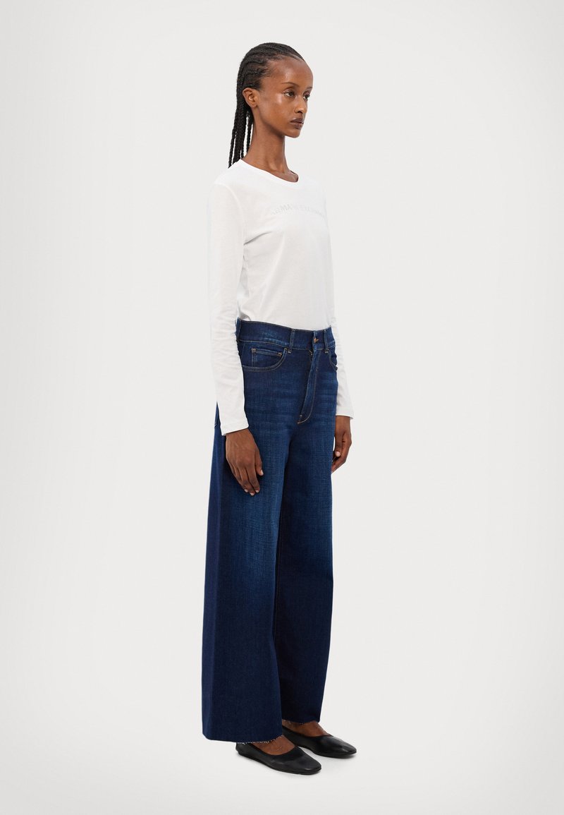 Camisa blanca de manga larga con cuello redondo, combinada con jeans de pierna ancha en azul oscuro. Unos zapatos planos negros completan el atuendo.