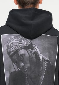 Μαύρη φούτερ με капюшон και μια μεγάλη μονόχρωμη γραφική απεικόνιση στην πλάτη, που απεικονίζει ένα κοντινό πορτρέτο ενός άνδρα, ντυμένου με ένα μοτίβο μαντήλι και αλυσίδες.