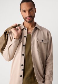 Bărbat cu păr scurt și barbă, purtând o jachetă bej cu nasturi peste un tricou verde mărgăritar, ținând o geantă din piele maro pe umăr, pe fundal alb.