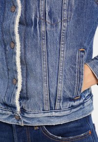 Giubbotto di denim blu con fodera in pile bianca, bottoni metallici, tasche cucite e una mano infilata nella tasca laterale destra.