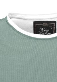 Primer plano del escote de una camisa de tela acanalada verde claro con un cuello interior blanco y una etiqueta de ropa negra dentro del cuello.