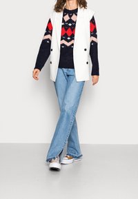 Blazer blanc sans manches porté sur un pull en tricot à motif bleu marine, rouge et rose, associé à un jean évasé bleu clair et des baskets blanches.
