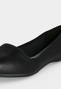 Chaussure plate noire à bout pointu avec une texture lisse et un talon bas, présentée sur un fond clair uni.