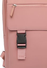 Rosa ryggsäck med en texturerad matt yta, ett svart plastspänne och en dragkedjeficka med en liten läderdragflik.