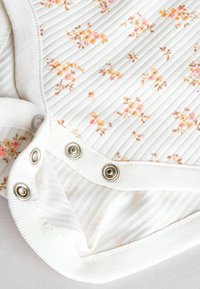 Weißes, blumenmusterndes Oberteil mit geripptem Textur, verziert mit kleinen rosa und gelben Blumen, Metallschnappverschlüssen am Boden.