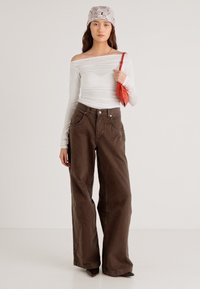 Witte off-shoulder top met lange mouwen, brede bruine broek, met een rode tas in de hand, en een lichtgeverfd hoofddoekje. Soepele stoftexturen.