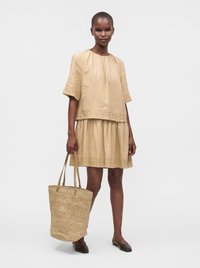 Femme portant une robe beige à manches courtes avec un ourlet brodé, tenant un sac cabas tissé, debout avec des chaussures plates marron sur un fond uni.
