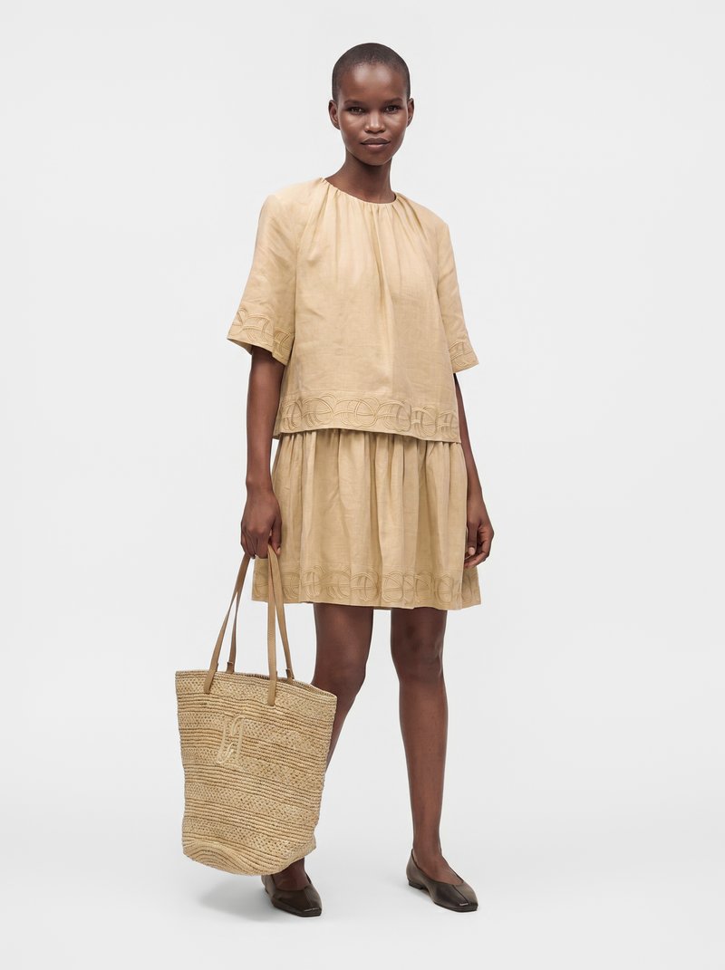 Femme portant une robe beige à manches courtes avec un ourlet brodé, tenant un sac cabas tissé, debout avec des chaussures plates marron sur un fond uni.