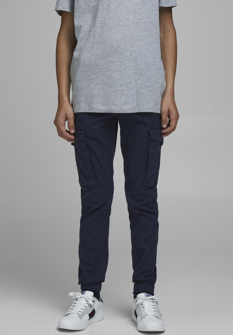 Personne portant un pantalon cargo marine, un t-shirt gris à manches courtes et des baskets blanches, debout devant un fond gris uni.