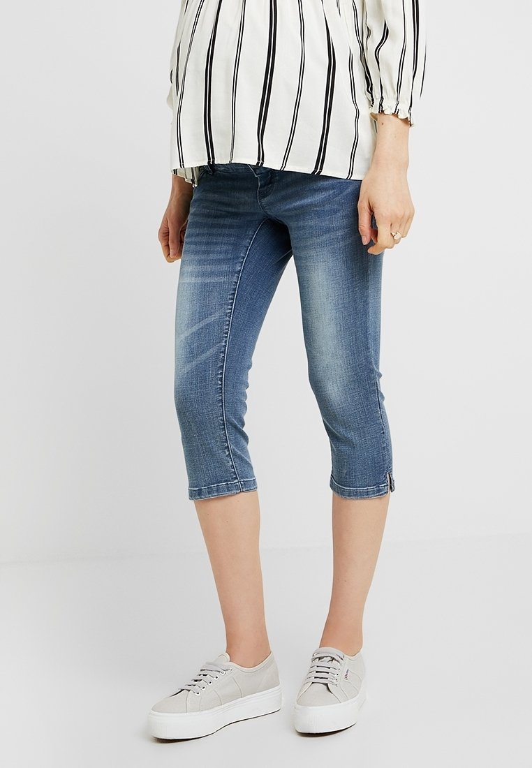 Denim capri-byxor, medelblå tvätt, slim fit, kort längd, med subtila blekningar, sidoknappsdetaljer, kombinerade med grå sneakers.