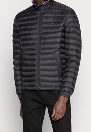 Uomo che indossa una giacca imbottita nera con cerniera e tasche laterali, con un piccolo logo rosso, bianco e blu sul petto, abbinata a pantaloni neri.