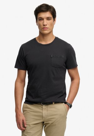 Młody mężczyzna w czarnej koszulce z krótkim rękawem i kieszonką na piersi oraz beżowych spodniach, stojący na tle jednolitej białej ściany.