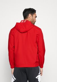 Rode waterdichte jas met een capuchon, elastische manchetten en een strak ontwerp. Glad textuur en minimalistische stijl, zicht van achteren.