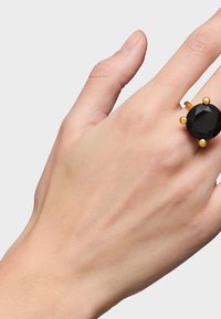 Anillo en tono dorado con una gran piedra negra facetada y acentos dorados redondos, mostrado en una mano contra un fondo sencillo.