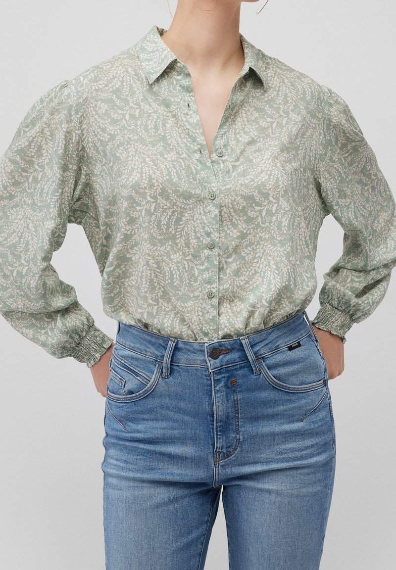 Femme portant un chemisier vert clair à manches longues avec un motif de feuilles blanches, rentré dans un jean bleu taille haute, les mains sur les hanches.