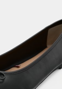 Ballerine en cuir noir avec un petit nœud sur le bout et une doublure intérieure beige, photographiée sur fond blanc.