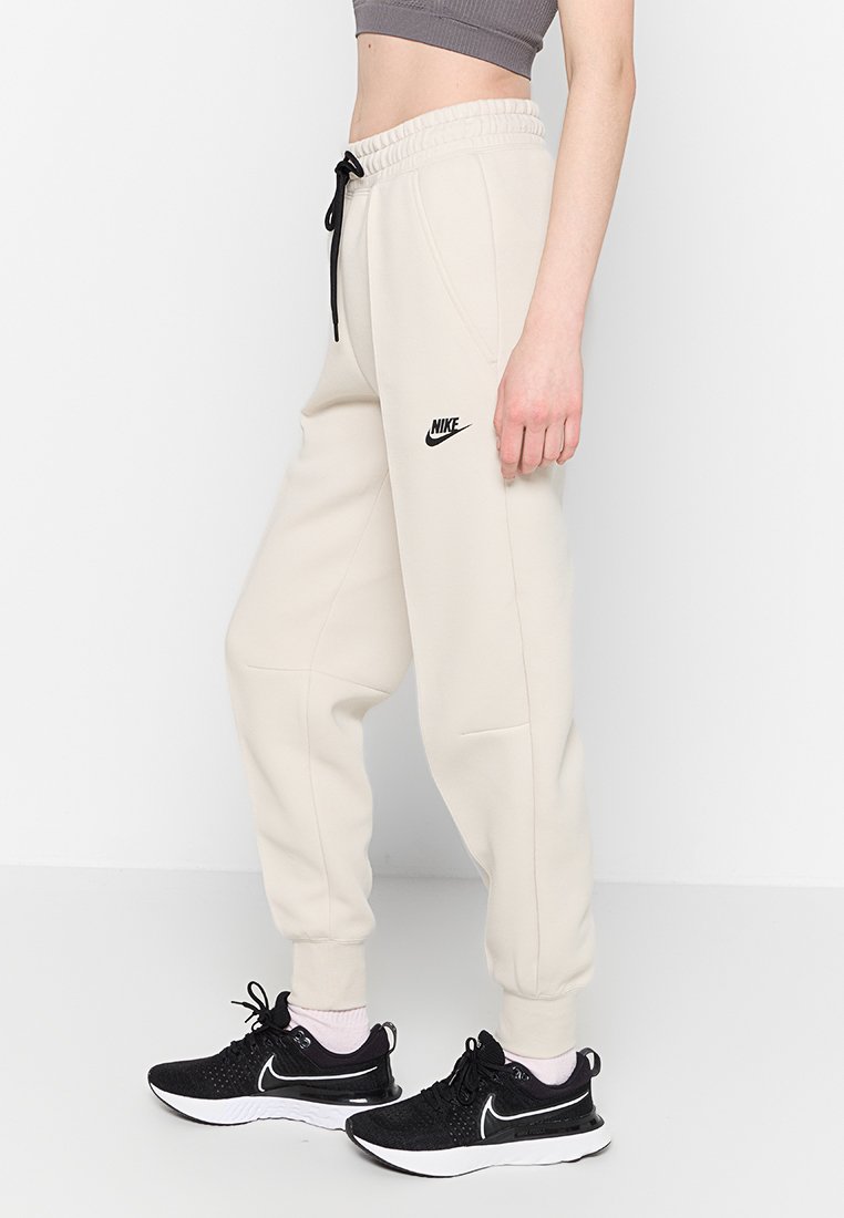 Pantalones de chándal beige con cintura elástica y cordón, bolsillos laterales, con un logo negro de Nike. Llevados con zapatillas deportivas negras y calcetines rosas.