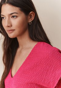 Haut rose texturé avec un col en V, associé à des boucles d'oreilles dorées de forme torsadée. Des cheveux lisses encadrent le visage.