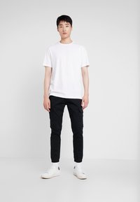 White short-sleeve t-shirt, black cargo pants with side pockets, white low-top sneakers, standing on a plain light background.