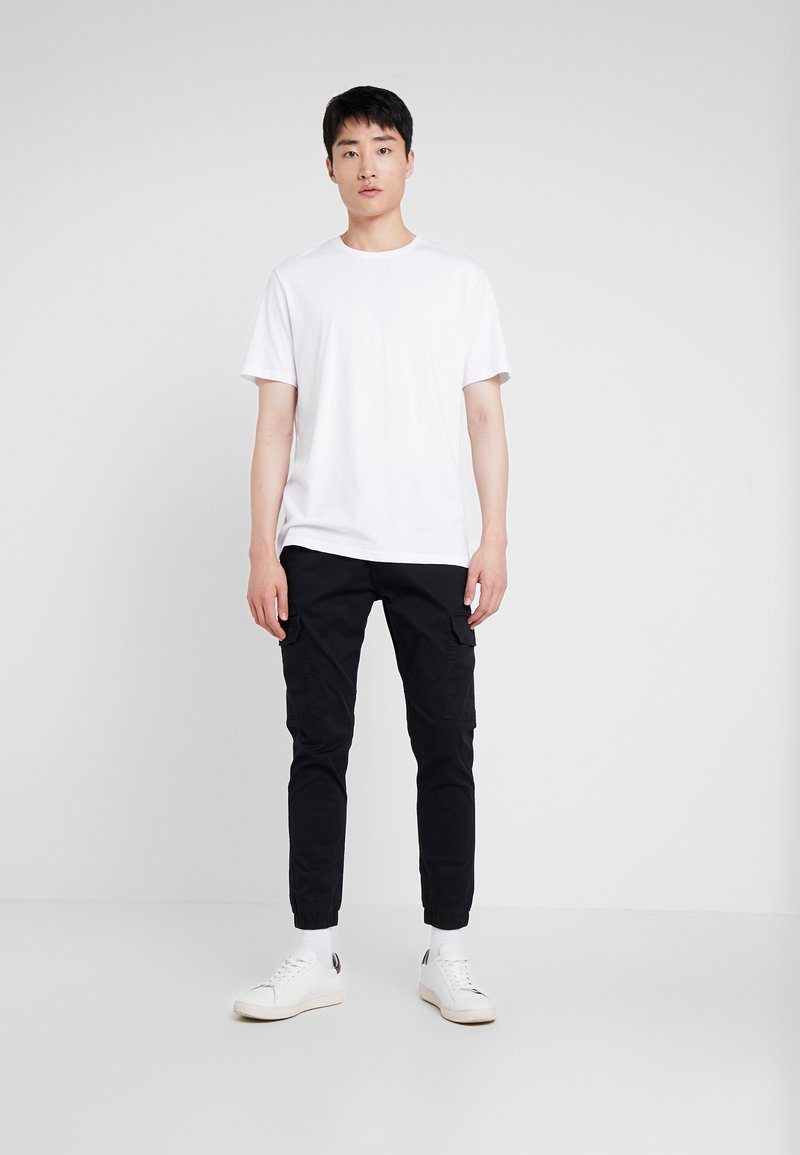 White short-sleeve t-shirt, black cargo pants with side pockets, white low-top sneakers, standing on a plain light background.