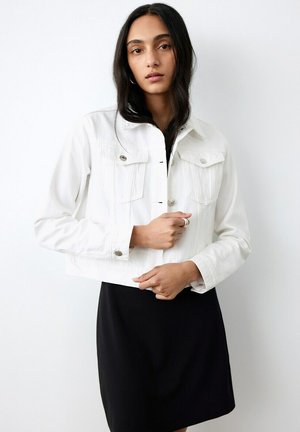 Mujer con cabello largo y oscuro, vestida con una chaqueta blanca abotonada sobre un vestido negro, posando con las manos entrelazadas a la altura de la cintura contra un fondo blanco.