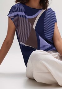 Kvinna klädd i en marinblå, lila och brun geometriskt mönstrad kortärmad topp med lösa vita byxor, sittande mot en ljus bakgrund.