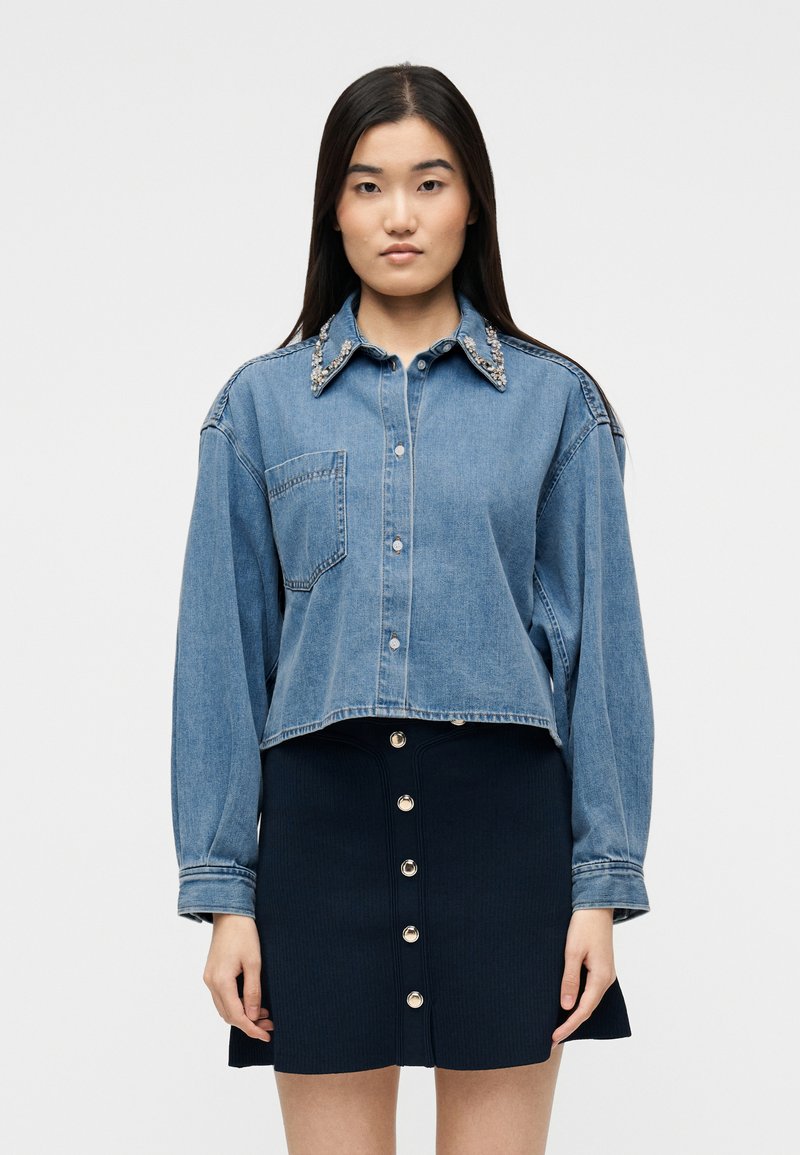 Νεαρή γυναίκα φοράει cropped τζιν μπουφάν με διακοσμητικό γιακά και μαύρη μίνι φούστα με κουμπιά, στέκεται μπροστά σε μονόχρωμο φόντο.