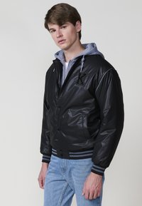 Giacca bomber nera lucida con cappuccio grigio, con polsini e orlo a righe, logo "K" ricamato sul petto e chiusura a bottoni.