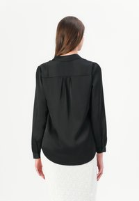 Black blouse with long sleeves, smooth texture, button cuffs, and a pleated back detail. Styled over a white textured skirt.