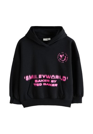 Ung kvinde iført en sort oversized hoodie med pink tekst og et smiley-logo, stående mod en ensfarvet lysegrå væg.