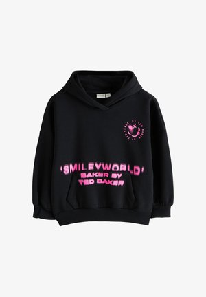 Giovane donna che indossa una felpa nera oversize con testo rosa e logo con faccina sorridente, in piedi contro un muro grigio chiaro uniforme.