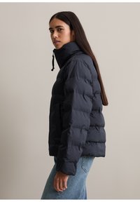 Doudoune marine à col haut, dotée d'une fermeture éclair et de détails élastiques. La veste a une texture matelassée et une coupe courte.