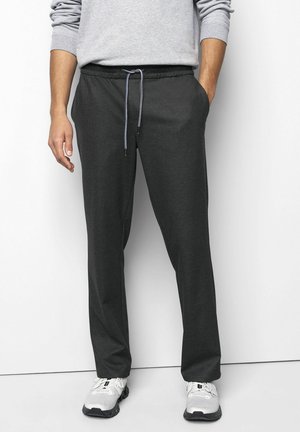 Man die donkere grijze joggingbroek met trekkoord en witte sportschoenen draagt, staand met één hand in zijn zak en zichtbare grijze sweatshirtmouwen.