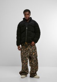 Mustast fliisist puffer-jope, millel on tõmblukuga sulgur, kombineeritud lahtiste, mustriliste rohelise ja pruuni tooniga cargo-pükstega. Neutraalset värvi tossud.