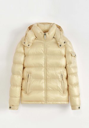 Veste matelassée beige avec fermeture éclair à l'avant, deux poches latérales zippées, poignets à boutons-pression, capuche amovible, et une poche à rabat sur la manche gauche.