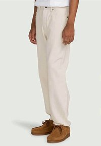 Béžové široké nohavice vyrobené z bavlny s predným zapínaním na gombík, dvoma bočnými vreckami a hladkou textúrou, skombinované s hnedými semišovými topánkami.