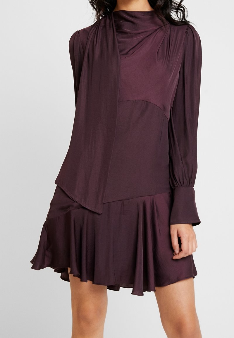 Une femme portant une robe longue à manches longues violet foncé avec un décolleté asymétrique et une jupe évasée plissée.