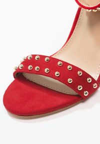 Sandalia de ante rojo con una banda ancha adornada con tachuelas doradas. Suela interior suave, diseño de punta abierta y base plana para mayor estabilidad.