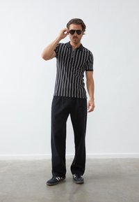 Polo shirt noir et blanc à rayures avec des manches courtes, associé à un pantalon noir ample et des baskets noires. Le modèle porte des lunettes de soleil.