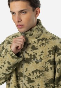 Fleecejakke i beige med grøn camouflage-mønster. Høj krave og fuld lynlås, med et logo på venstre bryst. Blød struktur.