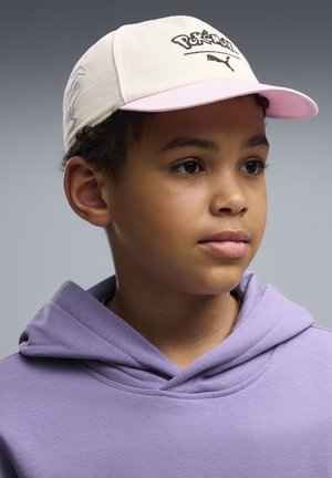 Młody chłopiec w beżowej i różowej czapce Pokémon Puma oraz jasnofioletowej bluzie z kapturem, patrzy lekko w prawo na szarym tle.