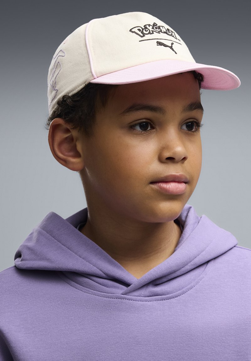 Jeune garçon portant une casquette beige et rose Pokémon Puma et un sweat à capuche violet clair, regardant légèrement vers la droite sur un fond gris.
