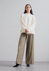 Kremowy, oversize sweter z dekoltem w kształcie litery V i elastycznym wykończeniem, zestawiony z szerokimi oliwkowymi spodniami. Modelka w czarnych szpilkach.