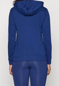 Osoba v jednoduché modré mikině a odpovídajících modrých legínách, viděná z pohledu zezadu na jednotným světlém pozadí.